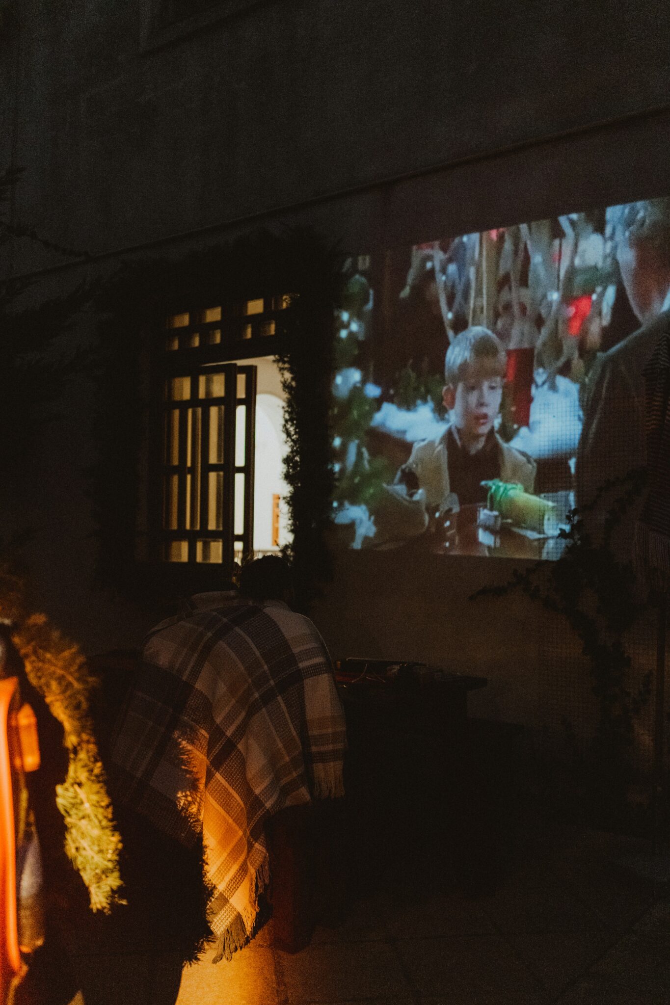 An outdoor scene showing a movie projected onto a wall, featuring a child actor, with a bench covered in blankets in the foreground.