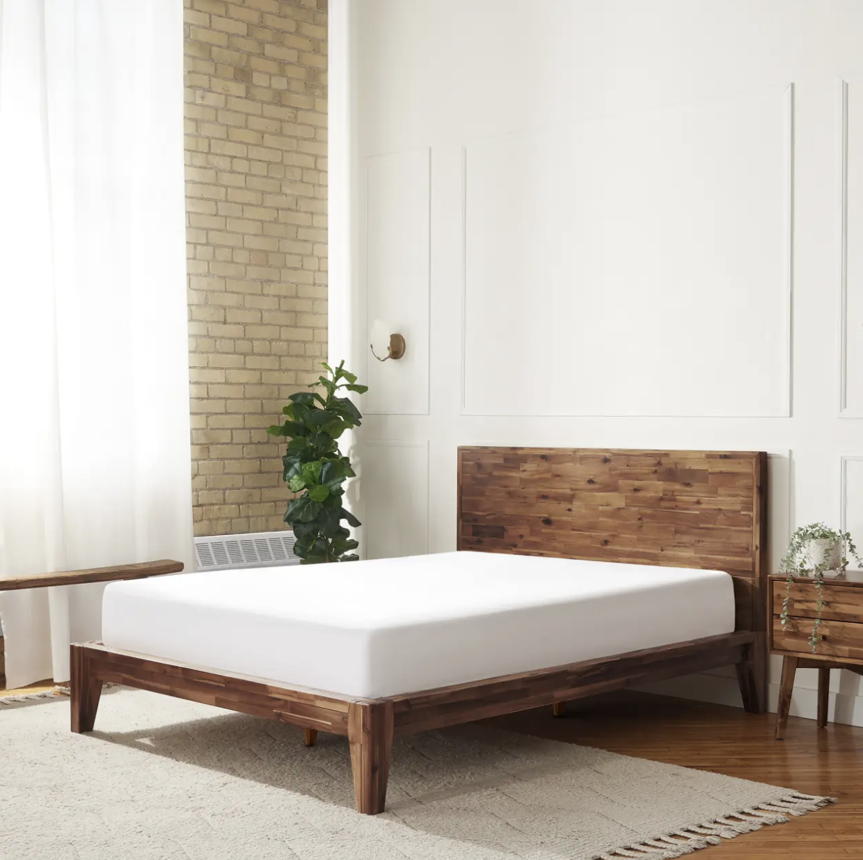A minimalist bedroom features a wooden bed frame with a white mattress, a nightstand, a potted plant, and natural light from large windows.