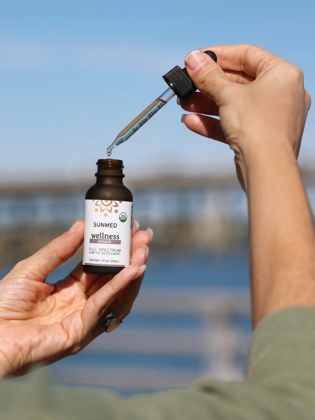 A person holds a dropper above a small brown bottle labeled "SUNMED Wellness Full Spectrum CBD Oil" with a blurred outdoor background.