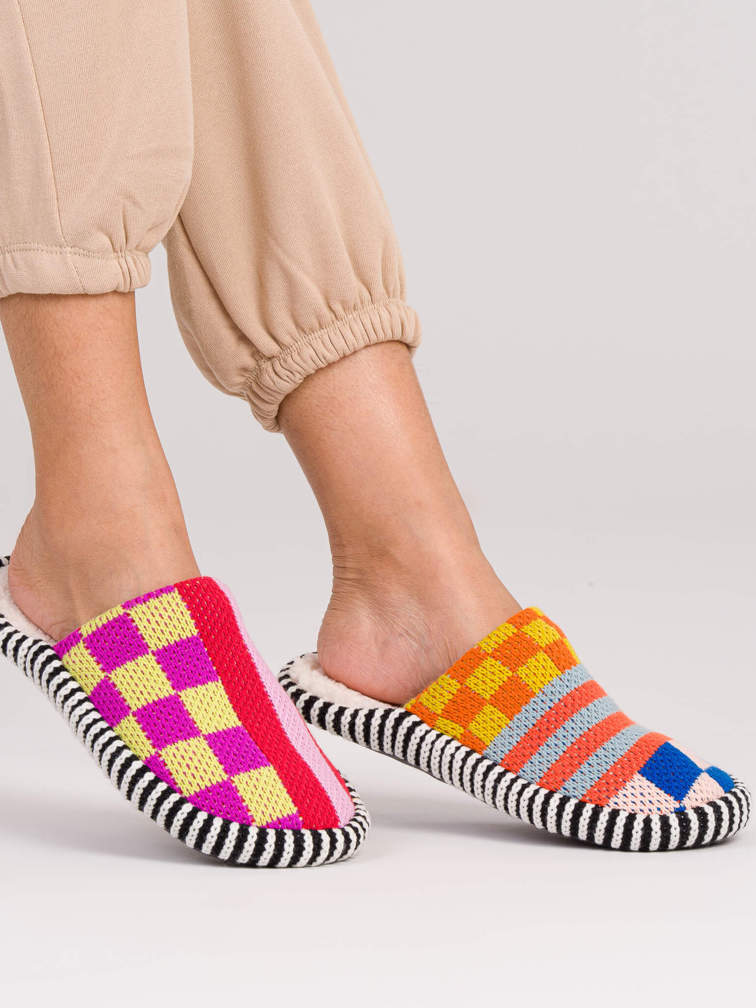 A person wearing mismatched, colorful slippers and beige cuffed pants stands on a light background.