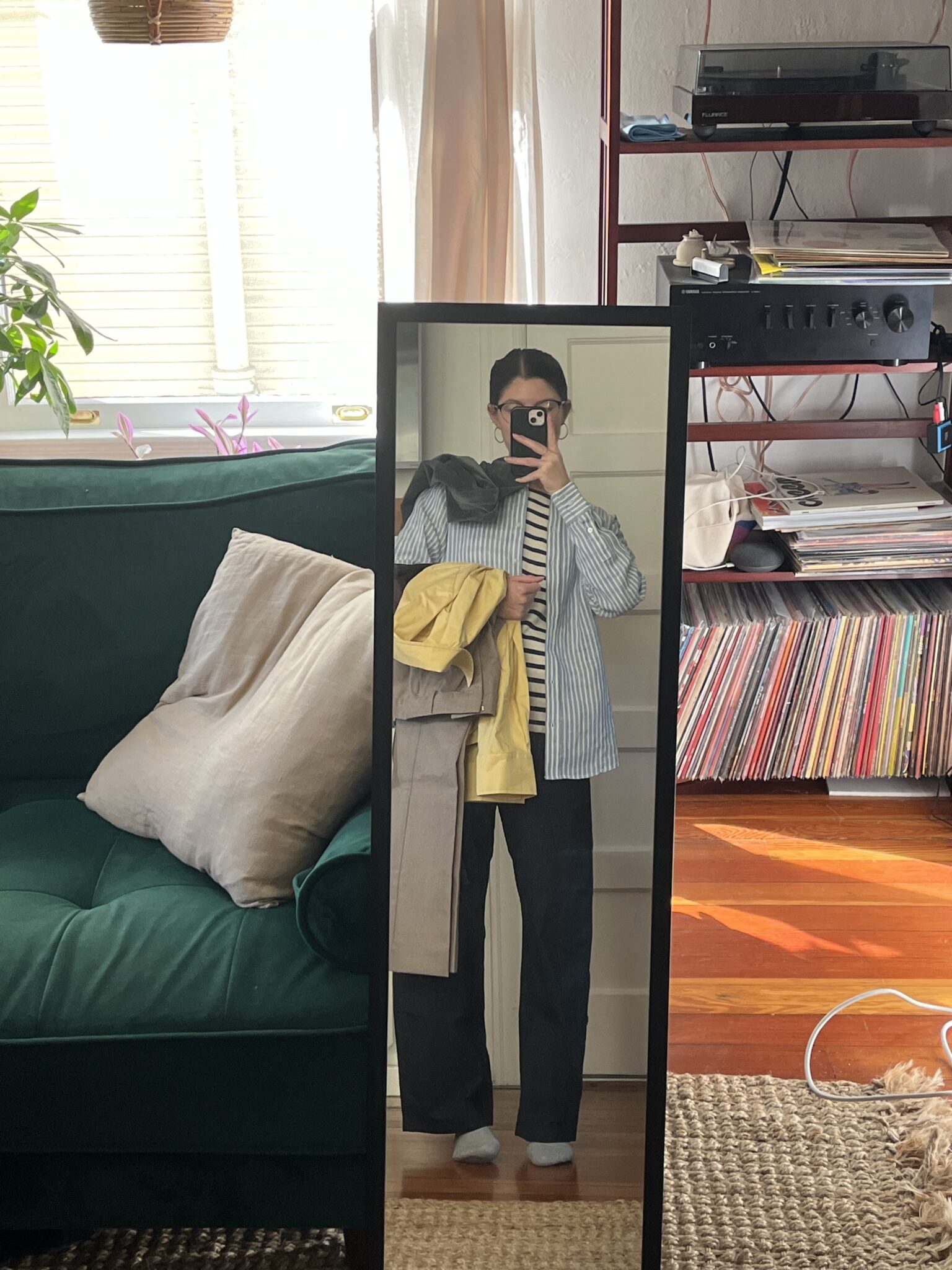 Person taking a mirror selfie in a living room, holding a yellow jacket and beige coat, wearing a striped shirt, blue overshirt, and dark pants. Shelves with records and electronics in background.