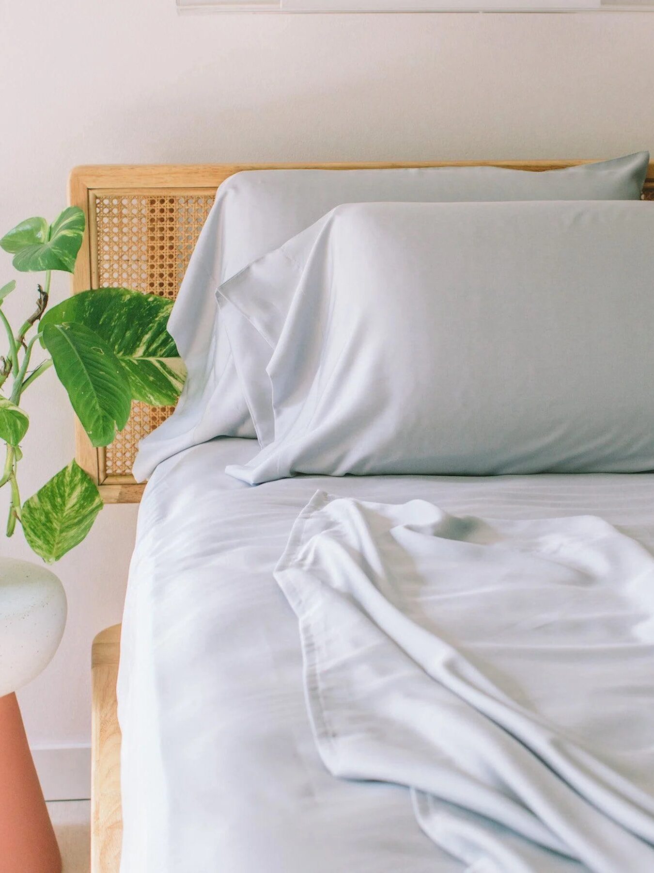 A neatly made bed with light gray sheets and pillows next to a small round table holding a potted green plant.