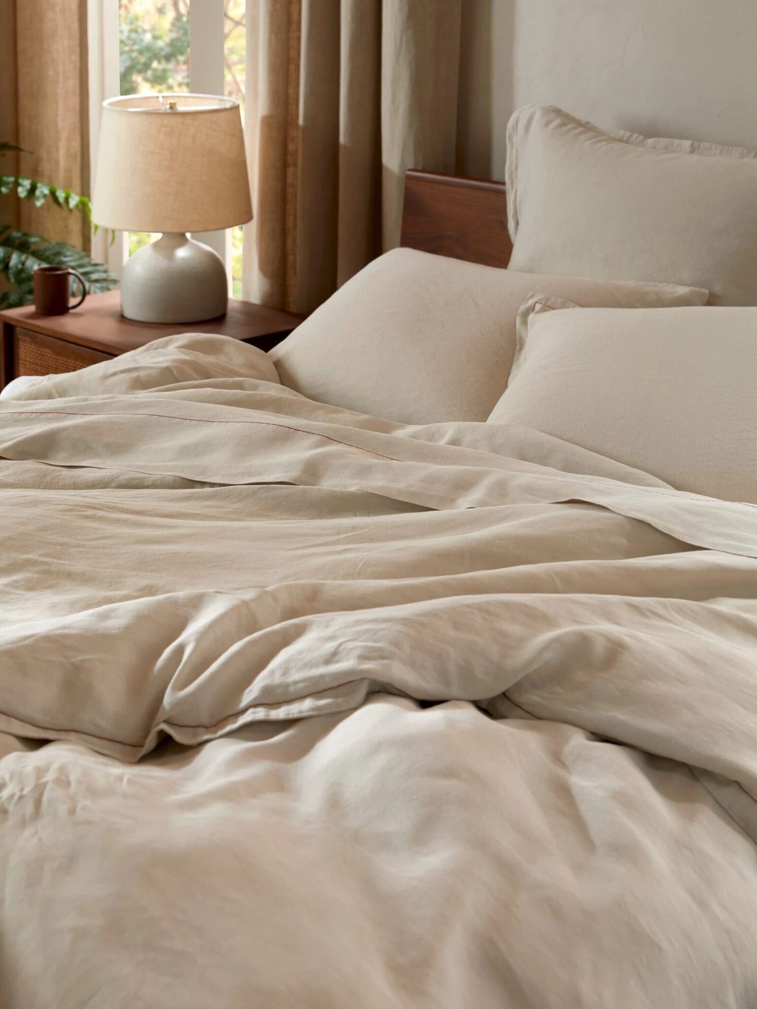 A neatly made bed with beige linens and pillows beside a wooden nightstand with a lamp, a mug, and a plant; sunlight filters through beige curtains.