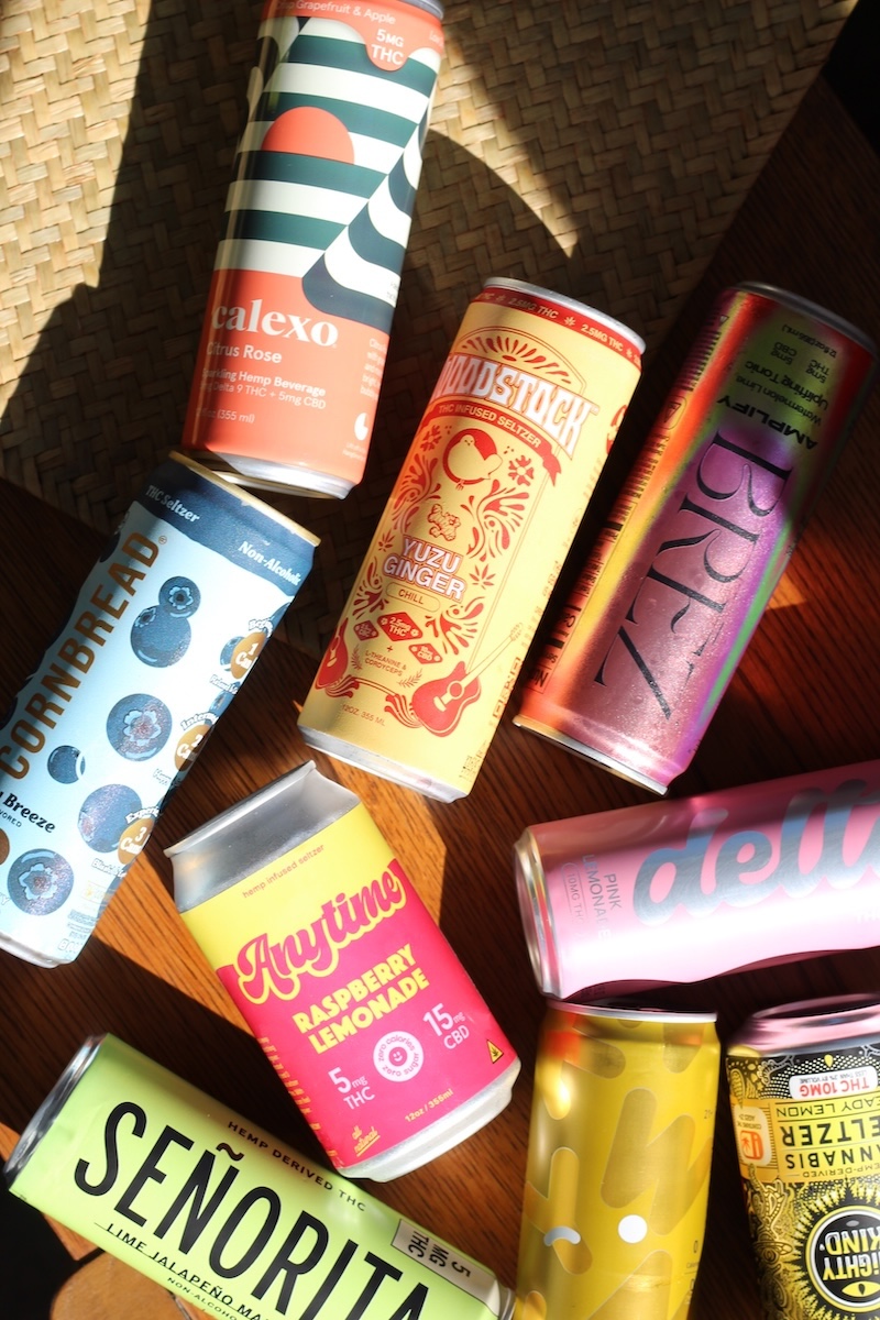 Nine colorful canned beverages, including brands like Calexo, Senorita, and Cornbread, displayed on a wooden surface with sunlight casting shadows.