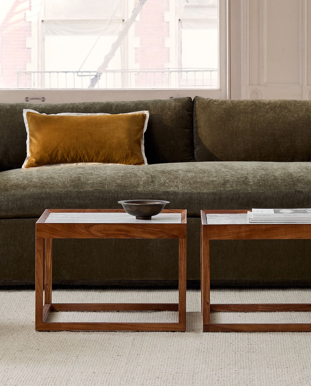 A green sofa from sustainable brands with a mustard yellow pillow sits behind two wooden coffee tables—one holding a shallow black bowl—in a room with a large window and beige carpet.