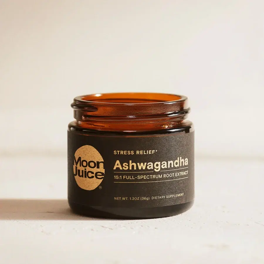 A brown glass jar labeled "Moon Juice Ashwagandha 15:1 Full-Spectrum Root Extract" sits on a light surface. The label notes it is a dietary supplement for stress relief.