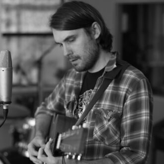 A man wearing a plaid shirt plays an acoustic guitar and sings into a microphone in a recording studio.