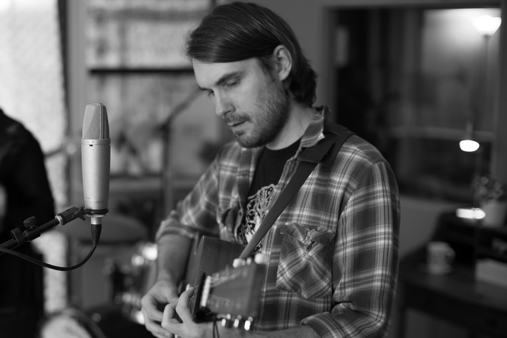 A man wearing a plaid shirt plays an acoustic guitar and sings into a microphone in a recording studio.