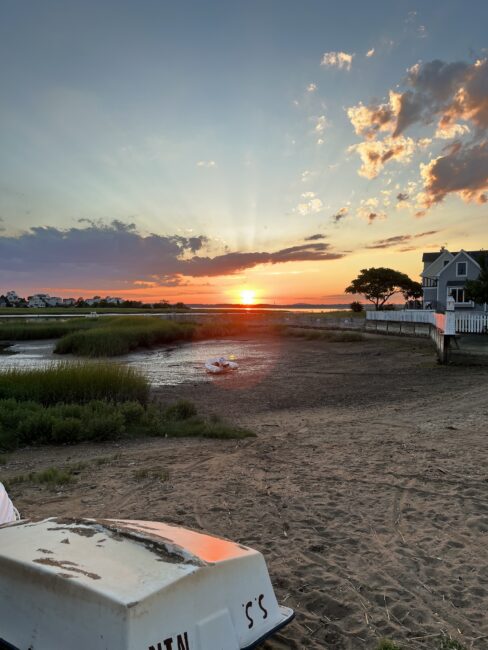 A sunset over a marsh with a sandy shore, grassy areas, a small boat, and houses in the background under a partly cloudy sky.