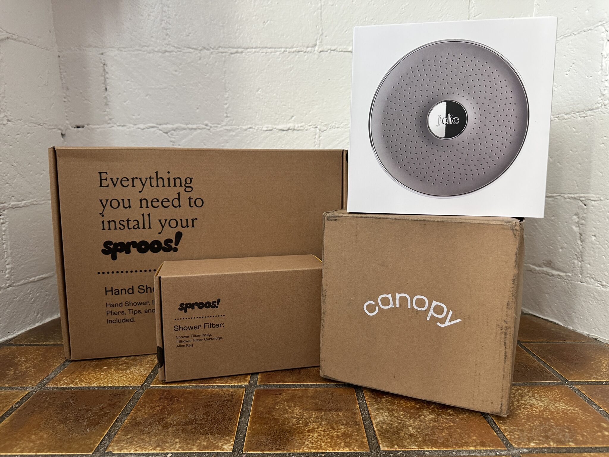 Four packaging boxes labeled "sproos!", "canopy", and "Jolie" are arranged on a tiled floor against a white brick wall.