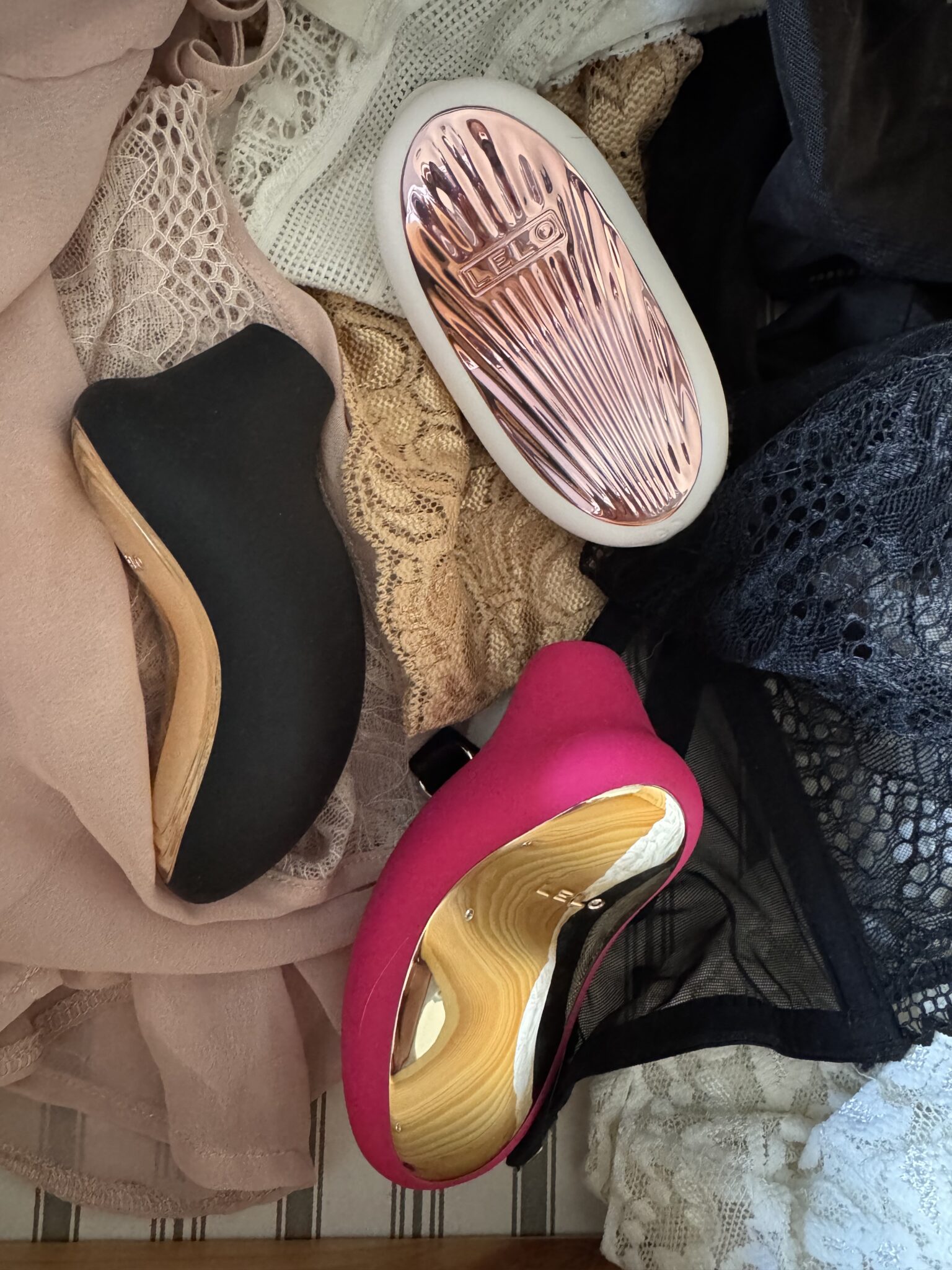 Two curved, wooden-handled items—one black, one pink—are placed on lace fabric, alongside a metallic rose gold oval object labeled "LELO.