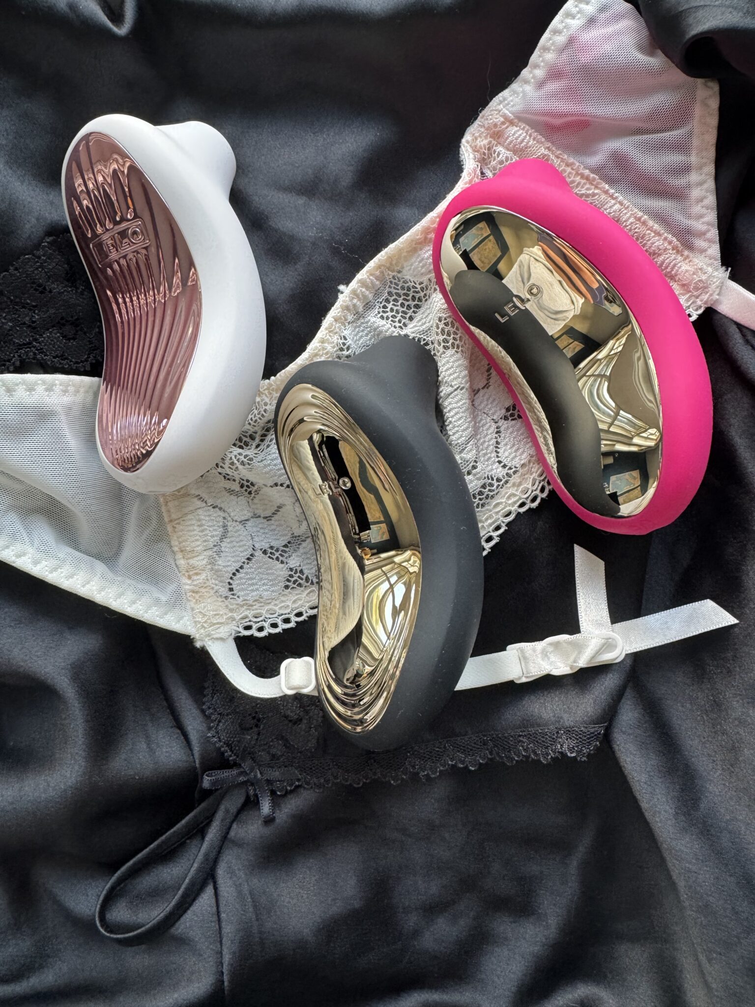 Three LELO personal massagers in white, black, and pink rest on a lace bra and black satin fabric.