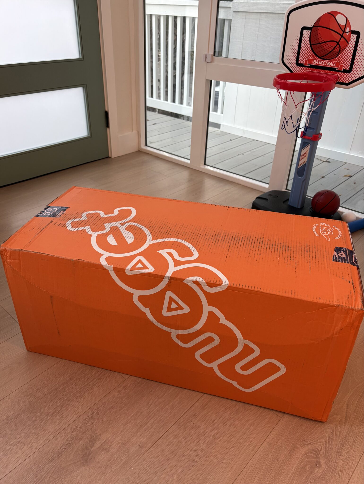 A large orange Nugget box sits on a wooden floor near a door and a children's basketball hoop.
