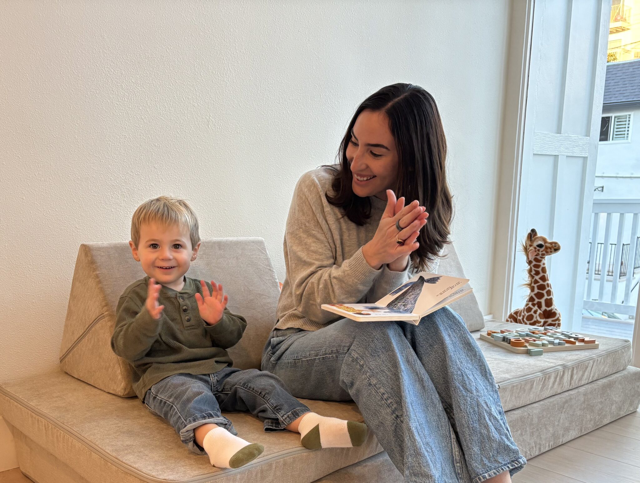 A woman reads a book to a young boy while they sit on a beige sofa; a toy giraffe and wooden puzzle are nearby.
