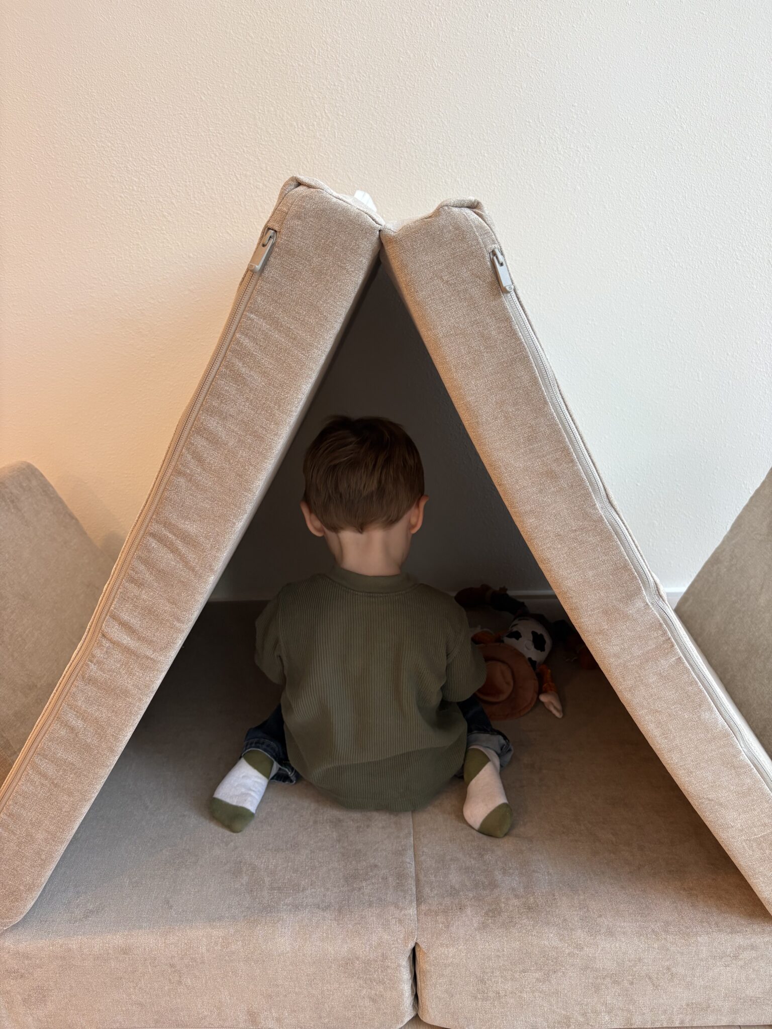 A young child sits with their back to the camera inside a makeshift tent made of beige couch cushions, playing with stuffed animals.