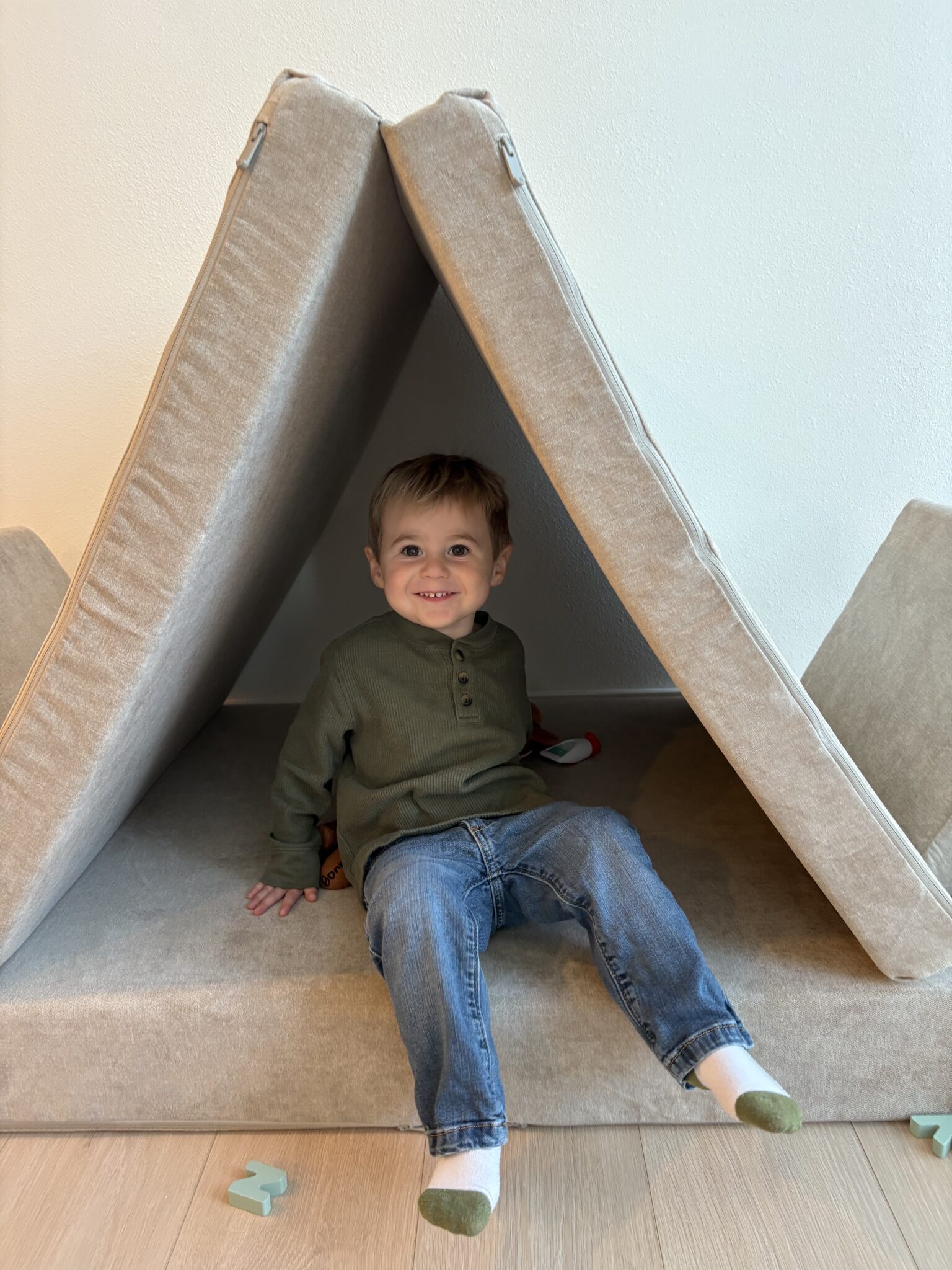 A young child sits smiling inside a small tent made from beige couch cushions, with puzzle pieces scattered on the floor nearby.