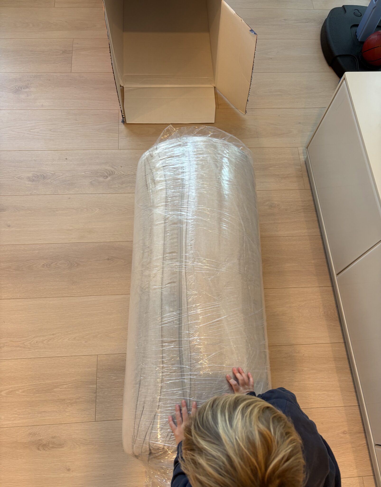 A child touches a rolled-up mattress wrapped in plastic on a wooden floor, with an open cardboard box nearby.