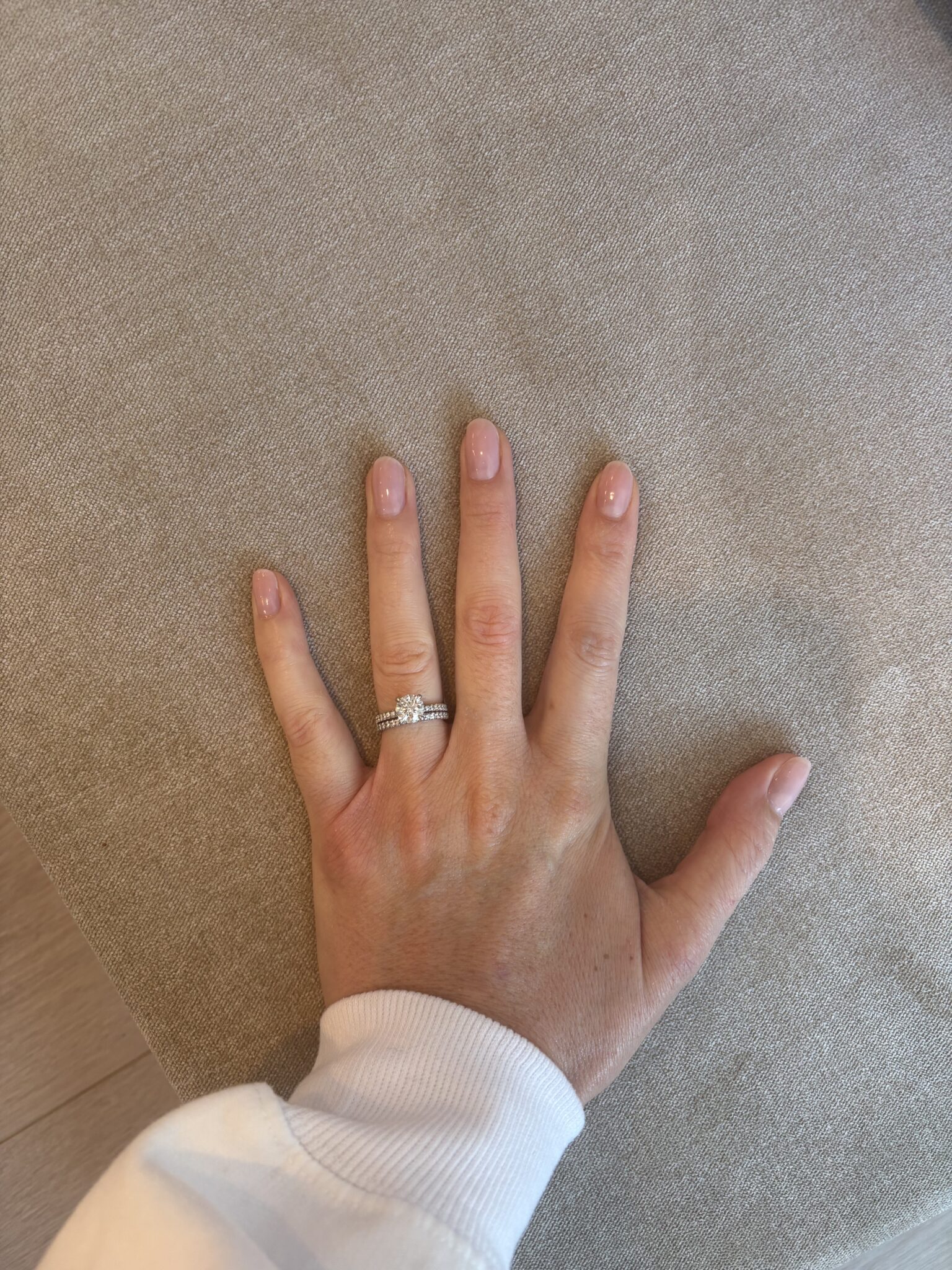 A left hand with a light pink manicure and two silver rings rests on a beige fabric surface.