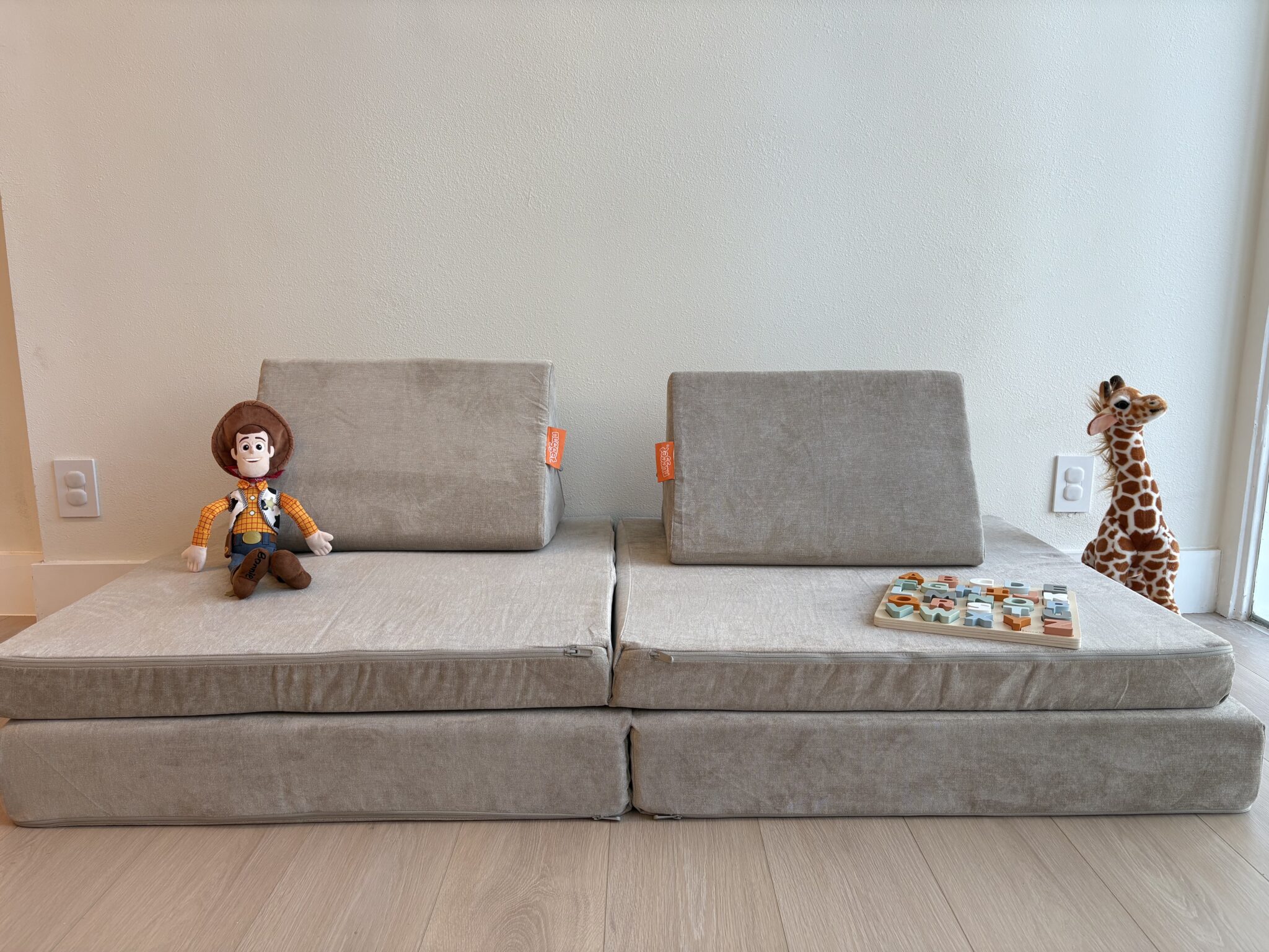 Two gray foam play couches with angled backrests, a Woody doll, a wooden animal puzzle, and a plush giraffe are arranged against a light-colored wall on a wooden floor.
