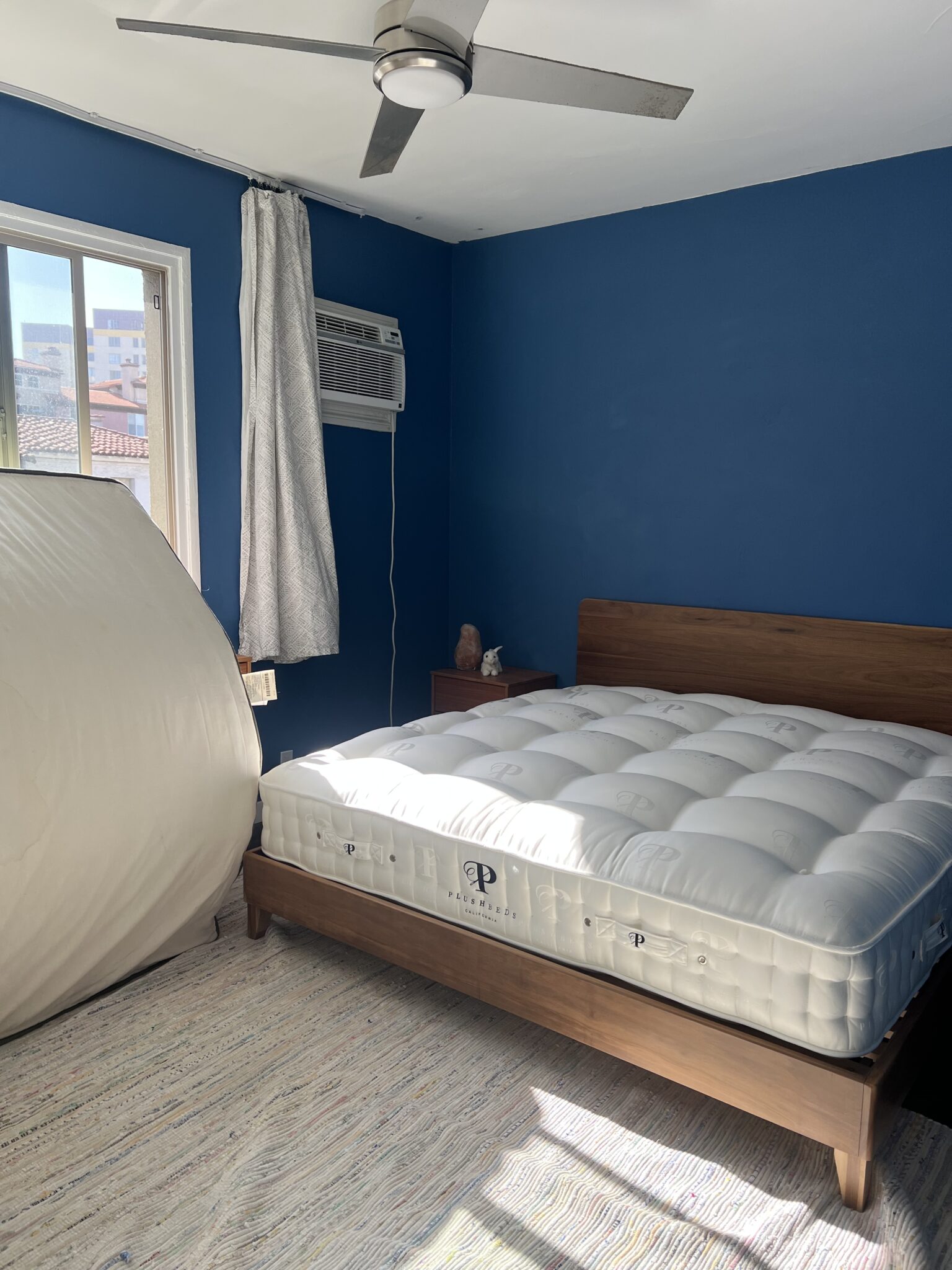 A neatly made bed with a thick white mattress sits in a blue-walled bedroom with a window, air conditioner, ceiling fan, and an upright mattress or foam object beside it.