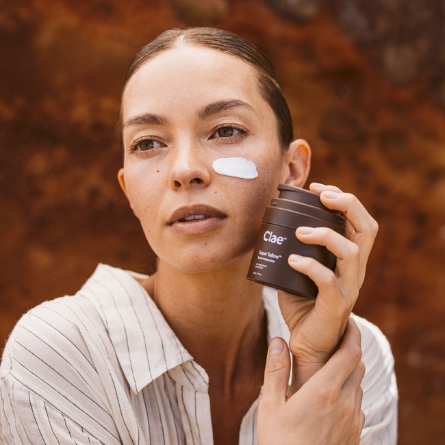 A person in a striped shirt applies white cream to their cheek while holding a brown bottle labeled "Clae" in front of a brown textured background.