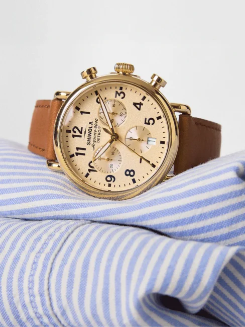 A gold chronograph wristwatch with a brown leather strap rests on a folded blue and white striped shirt.