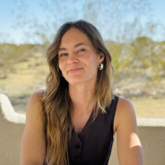A woman with long brown hair sits at an outdoor table holding a pen, with a notebook, coffee cup, and art supplies in front of her. Desert scenery is visible in the background.