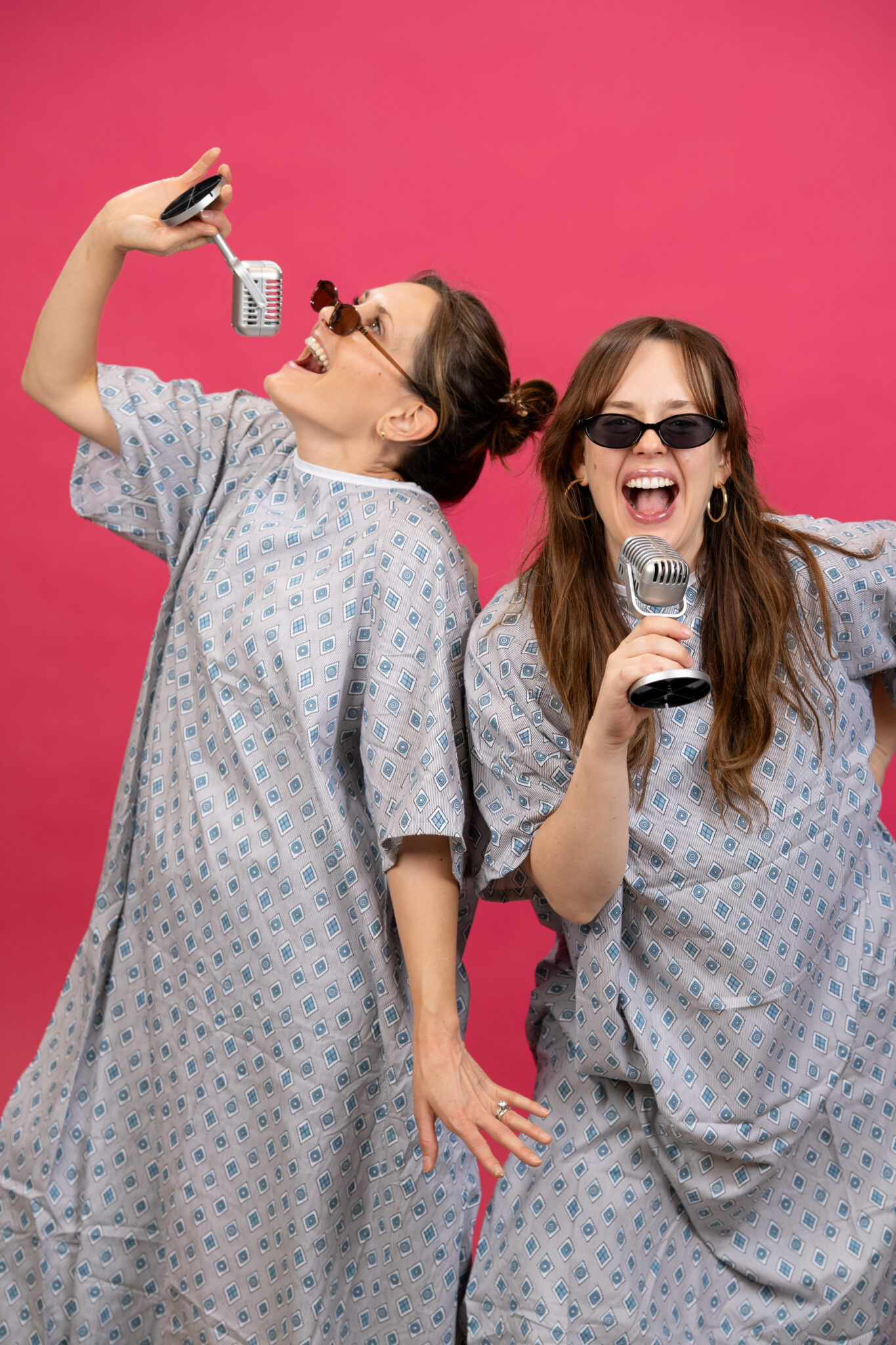 Two women in hospital gowns and sunglasses pose playfully, pretending to sing into retro microphones against a bright pink background.