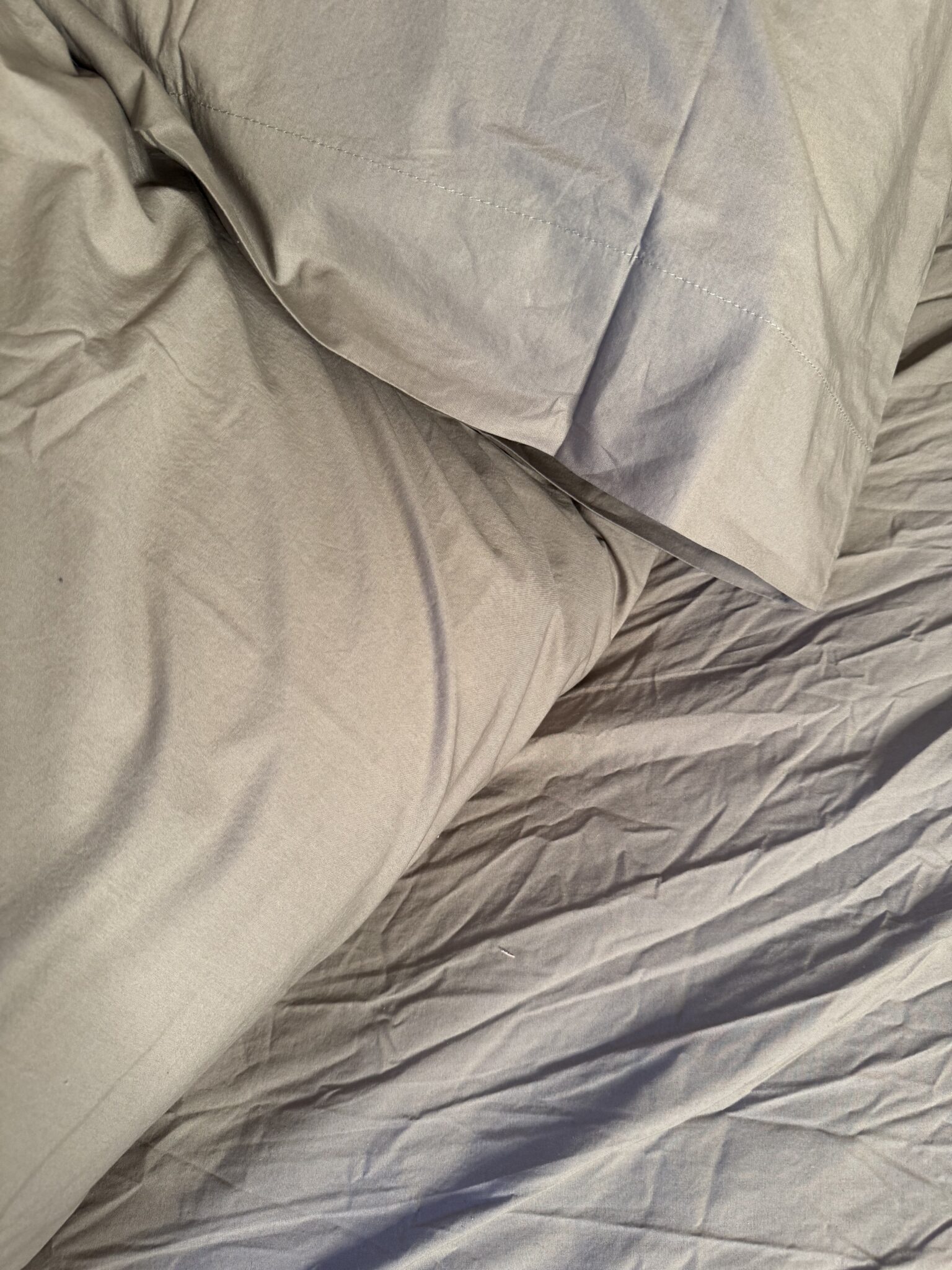 A close-up view of a wrinkled, light gray pillowcase and bedsheet on a bed.