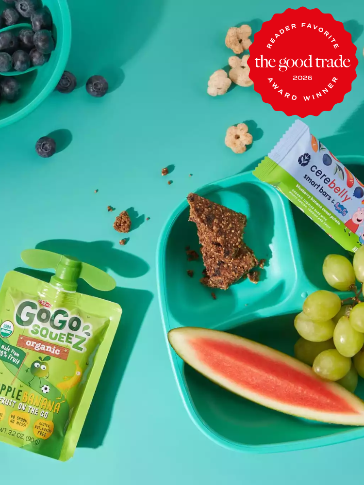A teal tray with grapes, a slice of watermelon, a snack bar, loose cereal pieces, blueberries, a fruit snack pouch, and a snack bar wrapper on a teal background.