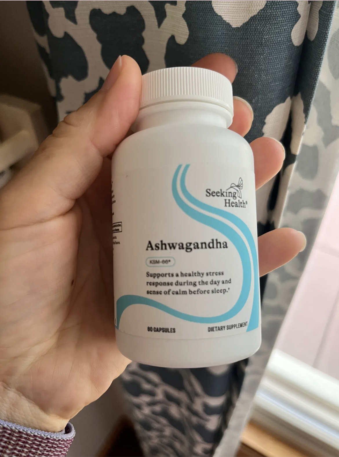 A hand holding a white bottle of Ashwagandha dietary supplement from Seeking Health, labeled to support stress response and calm before sleep.