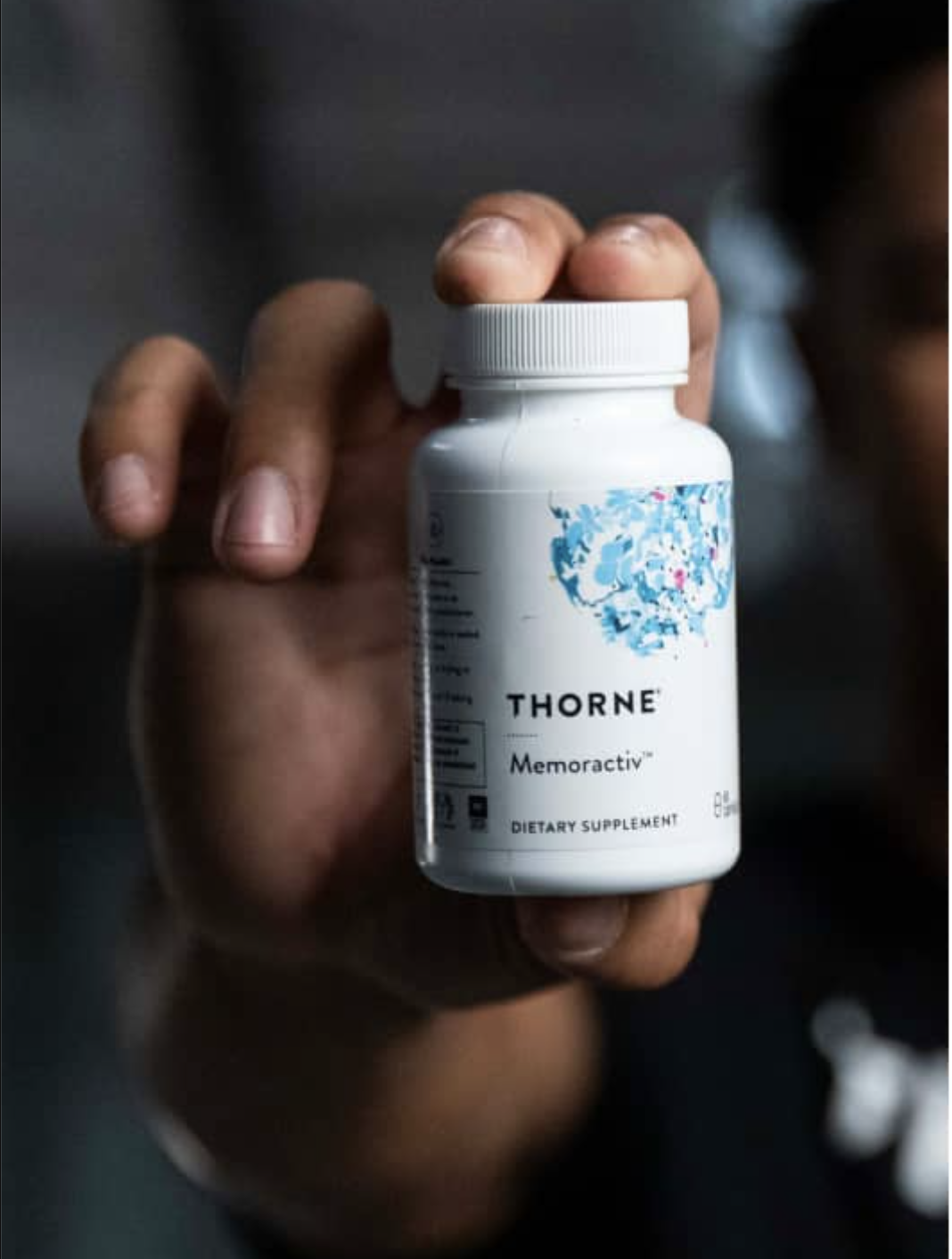 A person holds a white bottle of Thorne Memoractiv dietary supplement with a blue abstract design on the label.