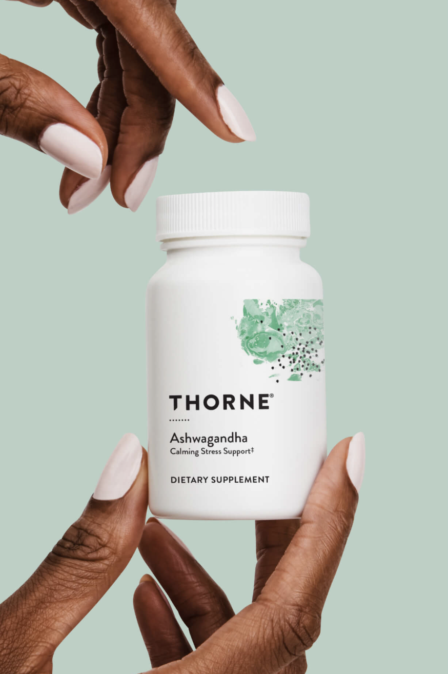 A person holds a bottle of Thorne Ashwagandha dietary supplement with a light green background.