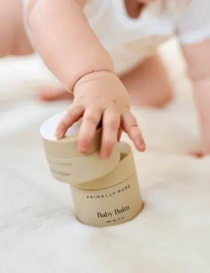A baby reaches for a container labeled "Primally Pure Baby Balm" on a soft, cream-colored surface.