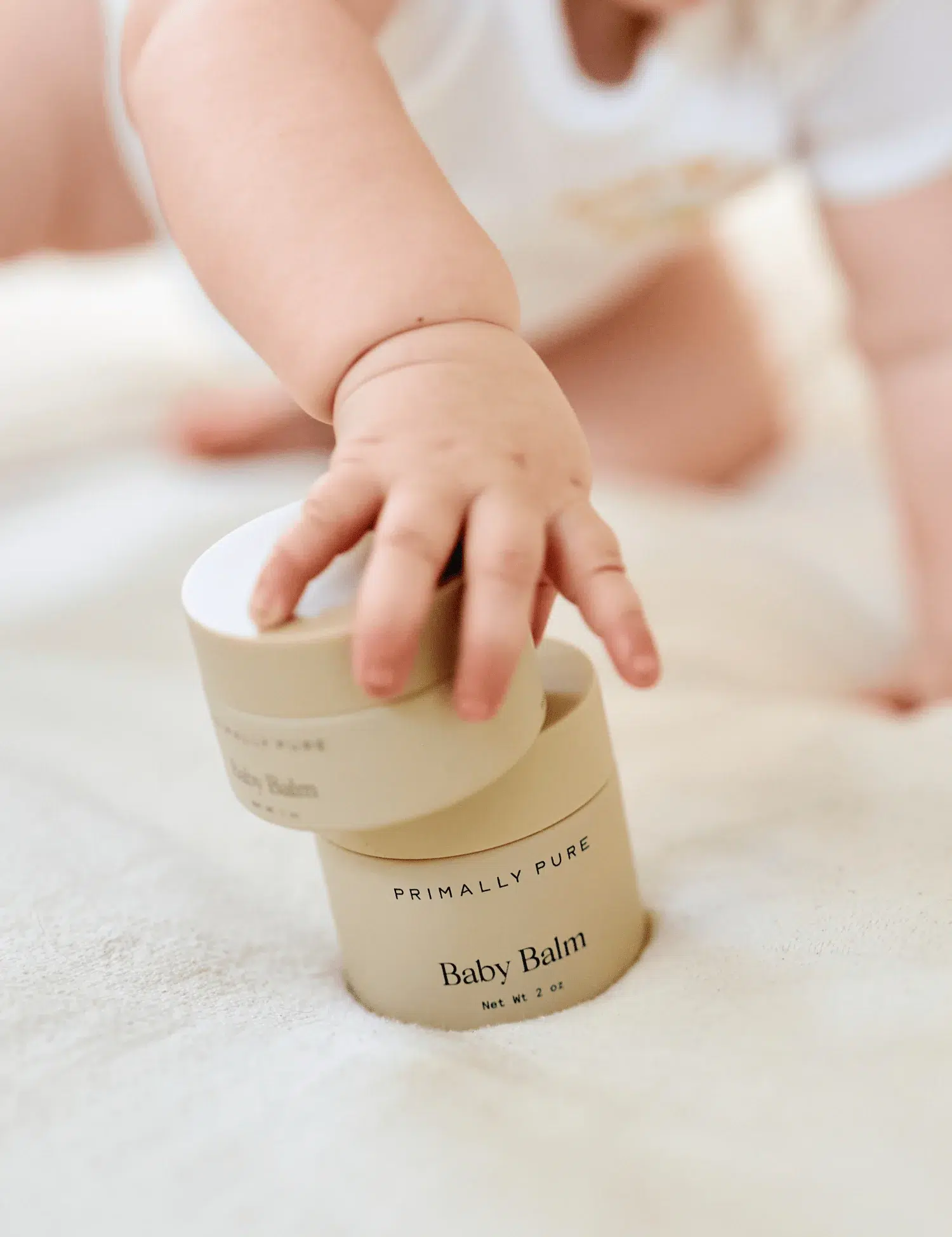 A baby reaches for a container labeled "Primally Pure Baby Balm" on a soft, cream-colored surface.