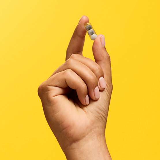 A hand with neatly manicured nails holds a two-toned capsule pill against a solid yellow background.