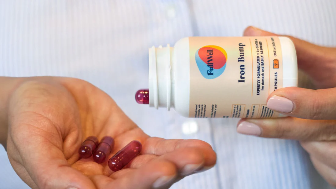 A person pours red iron supplement capsules labeled "Iron Bump" from a white bottle into their hand.