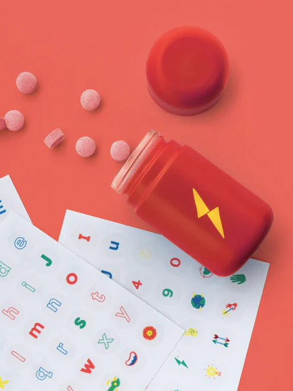 Three sheets of colorful alphabet stickers are displayed next to a red jar with a yellow lightning bolt and scattered pink tablets on a red surface. A “Free Sticker Pack” label is shown.