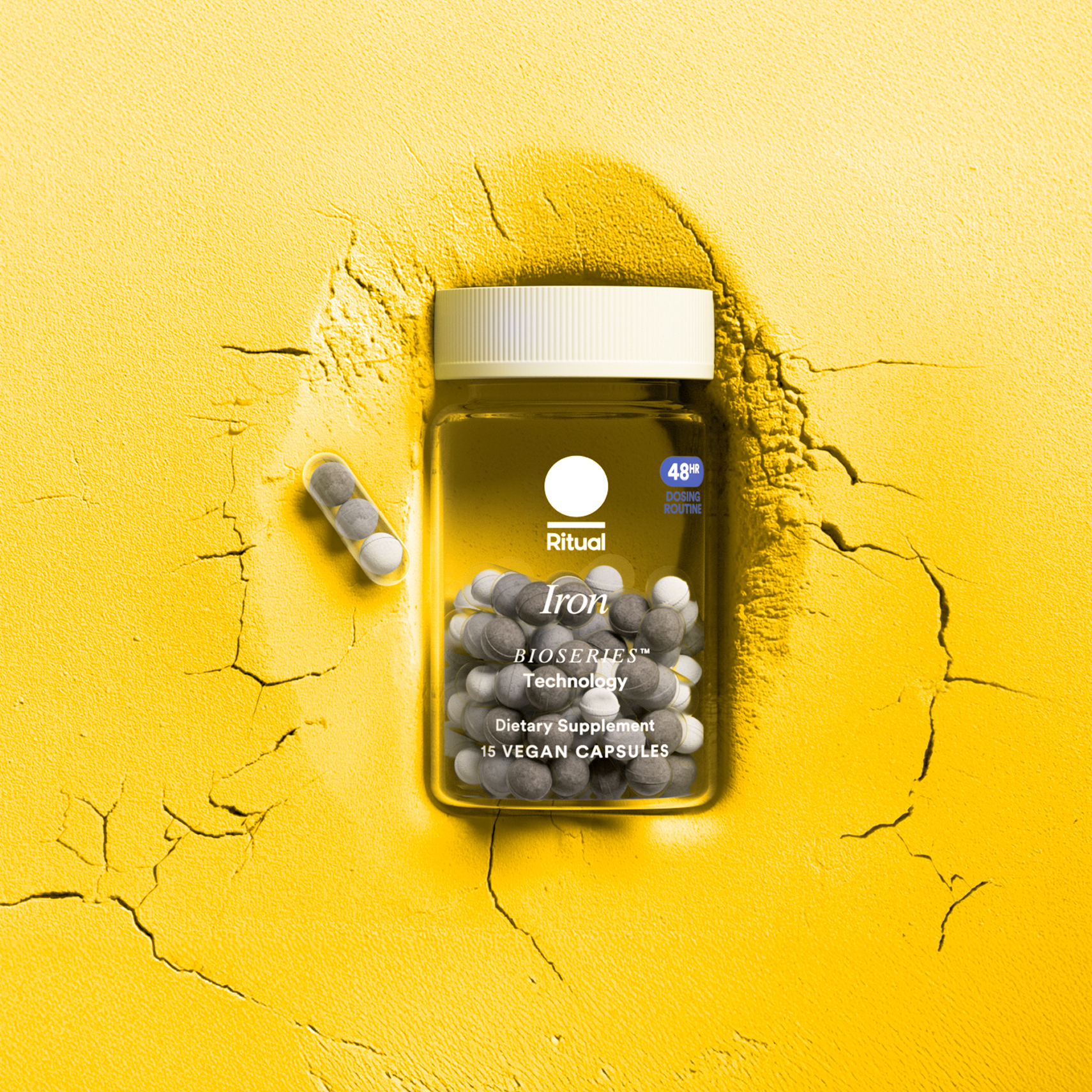 A jar of Ritual Iron dietary supplement capsules sits on a cracked yellow surface, with two capsules placed beside the jar.
