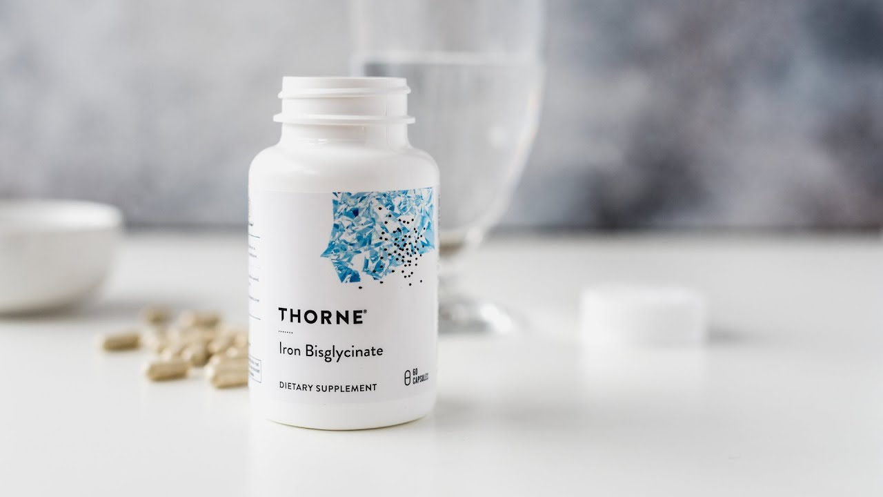 A white bottle labeled "Thorne Iron Bisglycinate" dietary supplement sits on a white surface next to a glass of water, pills, and a white bottle cap.