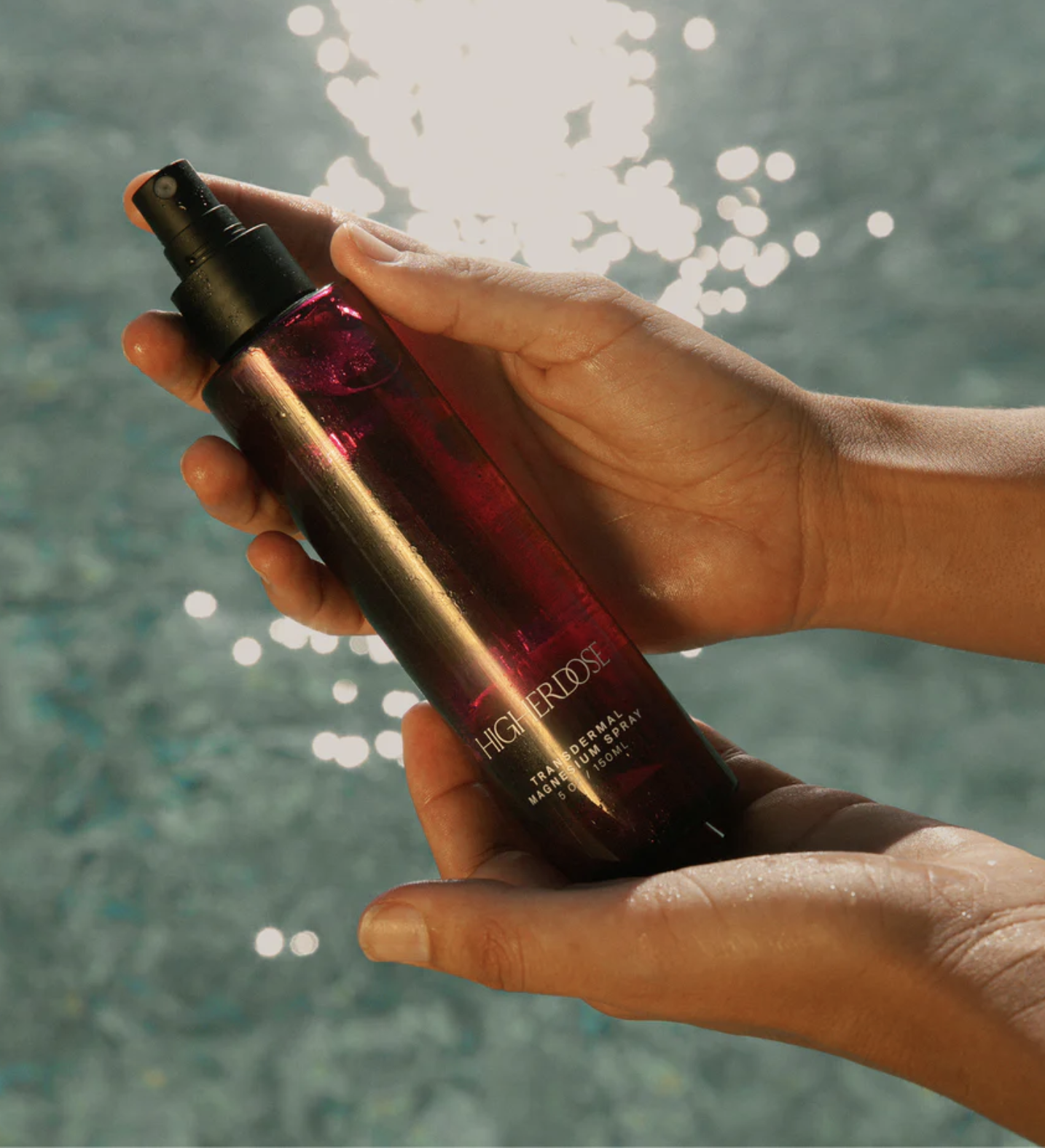 A person holds a maroon spray bottle with a black nozzle in front of a sunlit, rippling water background.