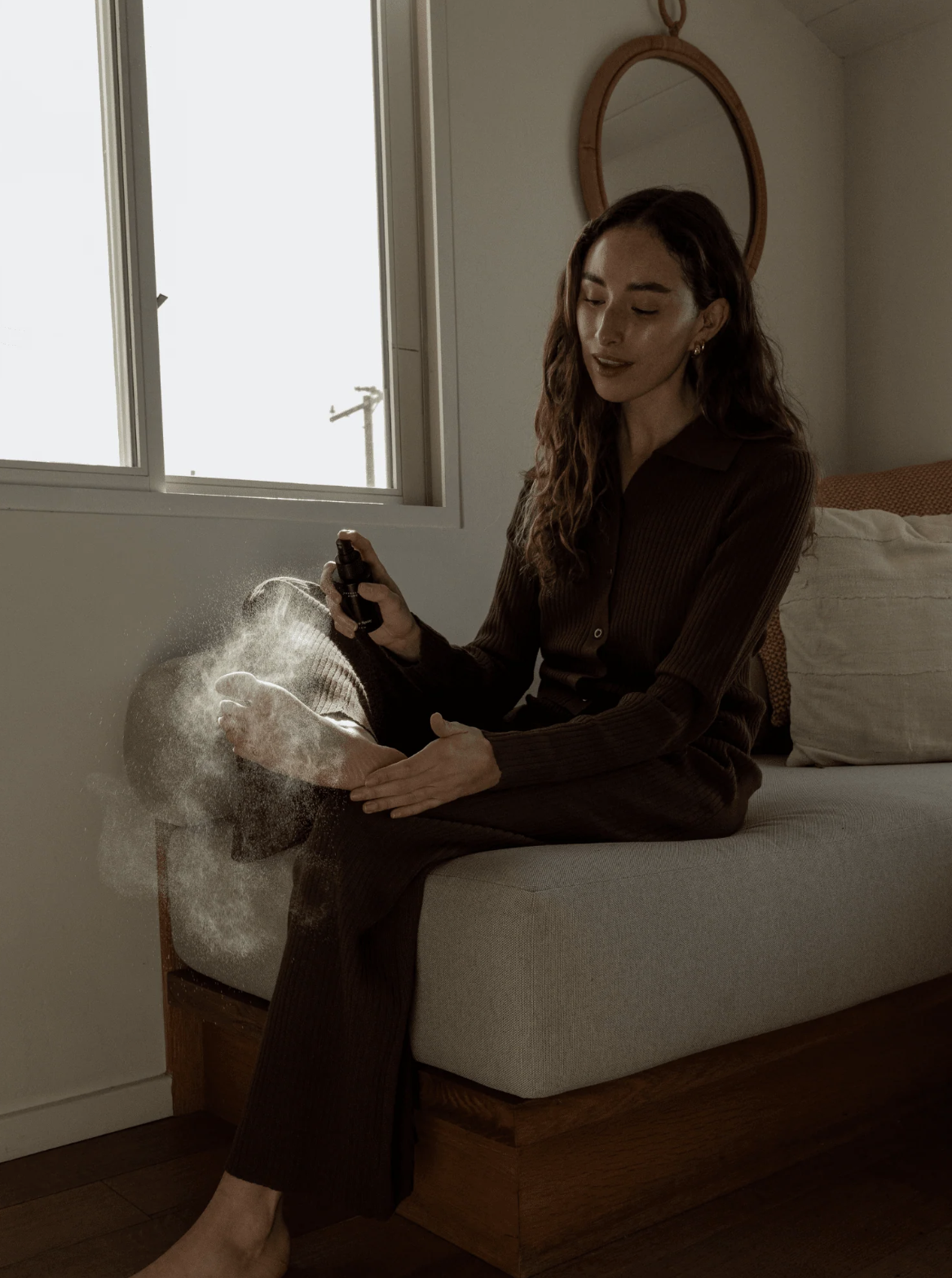 A woman sitting on a bed sprays a substance onto her bare foot, creating a visible mist in the air.