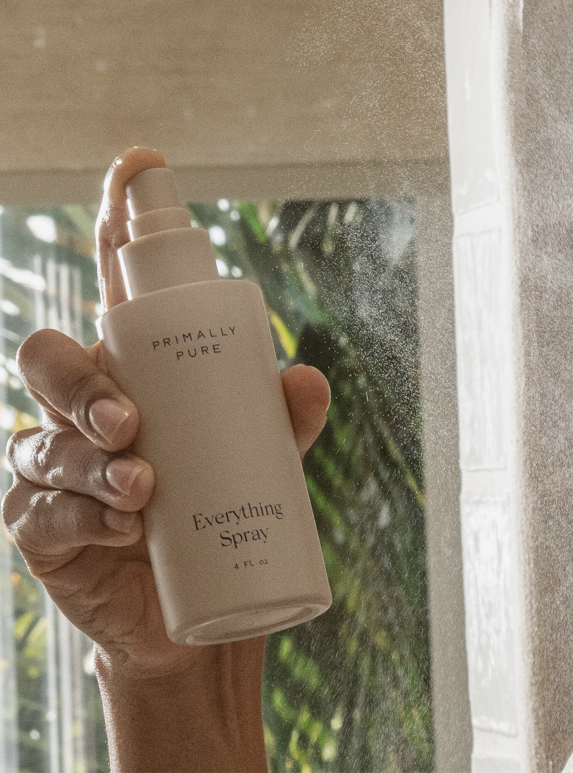 A hand holds and sprays a bottle labeled "Primally Pure Everything Spray" in front of a window with sunlight and greenery visible in the background.