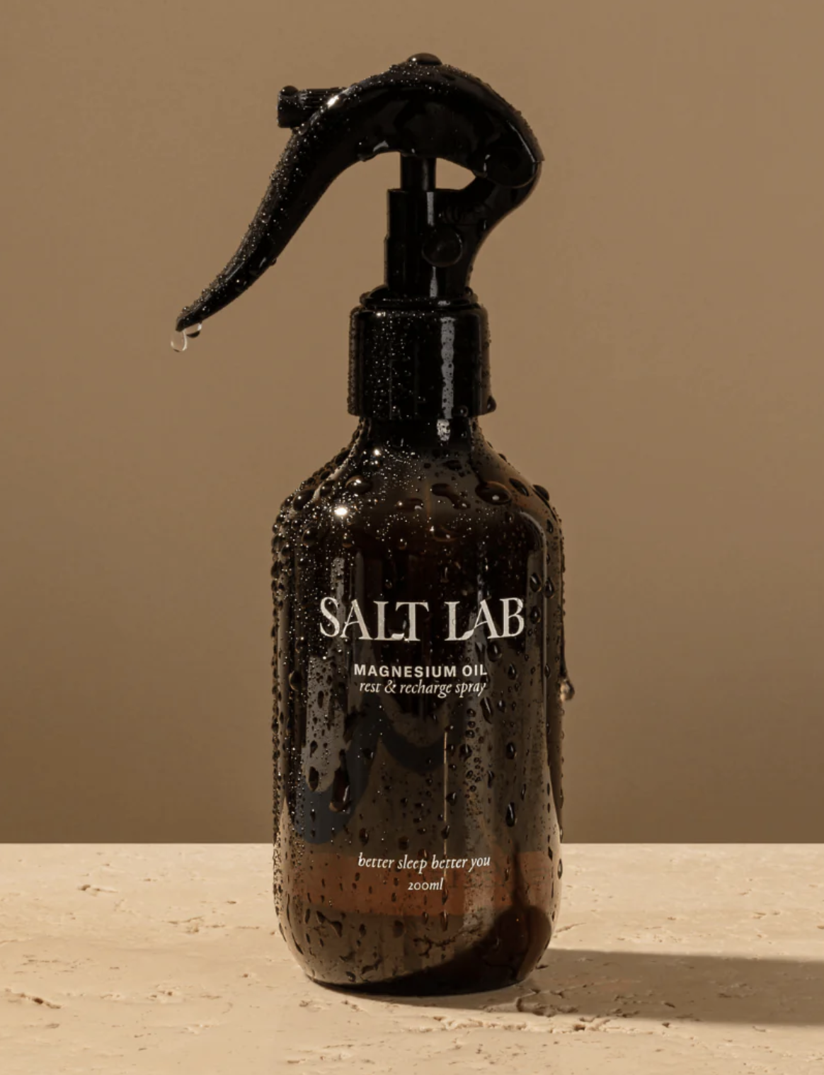 A brown glass spray bottle labeled "Salt Lab Magnesium Oil rest & recharge spray" with water droplets on it, placed on a light surface against a beige background.
