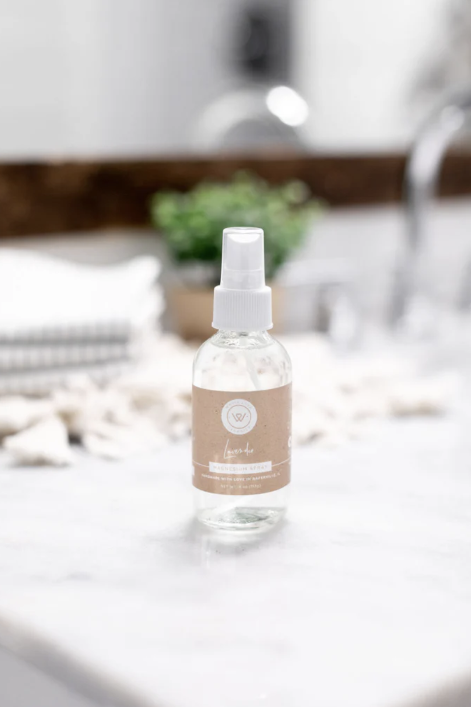 A clear spray bottle with a beige label sits on a marble bathroom countertop, with blurred towels, decor, and a plant in the background.