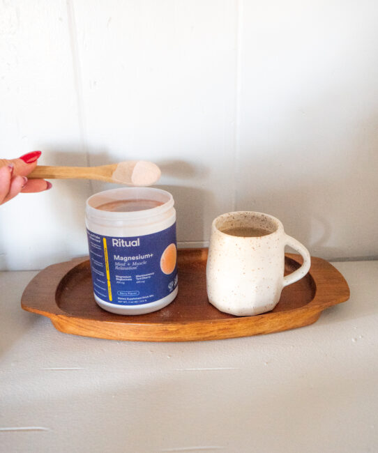 A hand scoops powder from a Ritual Magnesium+ container next to a ceramic mug on a wooden tray.