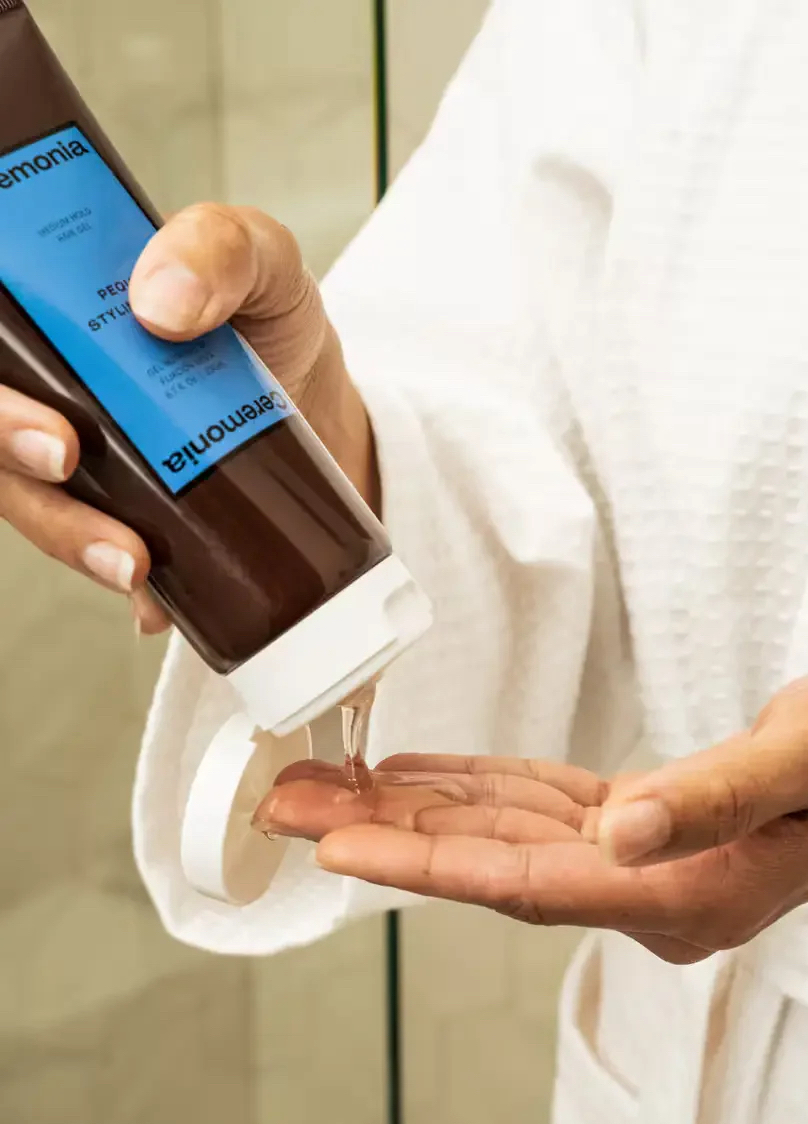 A person in a white robe dispenses clear liquid from a brown bottle with a blue label onto their palm.