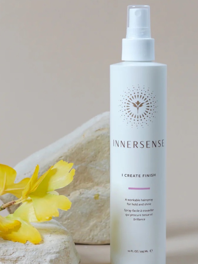 A white bottle of Innersense "I Create Finish" hairspray stands upright on a light surface, with yellow flowers and beige rocks in the background.