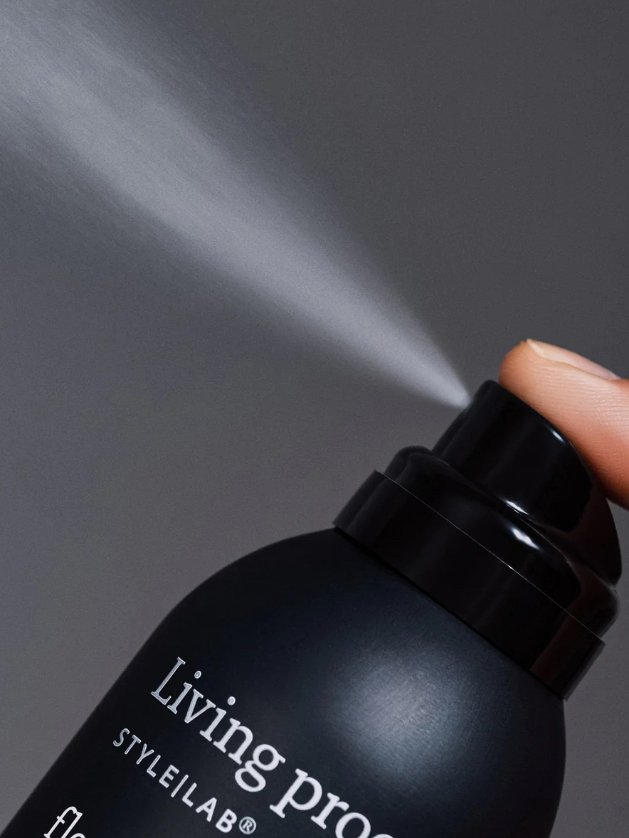 A close-up of a hand pressing a black spray can labeled "Living proof," releasing a fine mist against a neutral gray background.