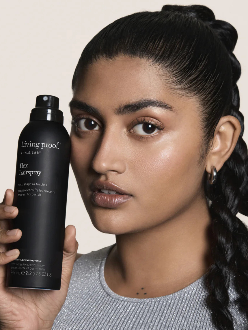 A woman with a long braid holds a can of Living Proof Style Lab Flex Hairspray near her face, looking at the camera.
